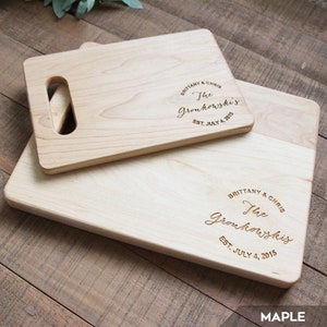 May include: Two wooden cutting boards with a personalized message. The top board has a handle and the bottom board is larger. The message reads "Brittany & Chris The Gronkowski's Est. July 4, 2015". The boards are made of maple wood.