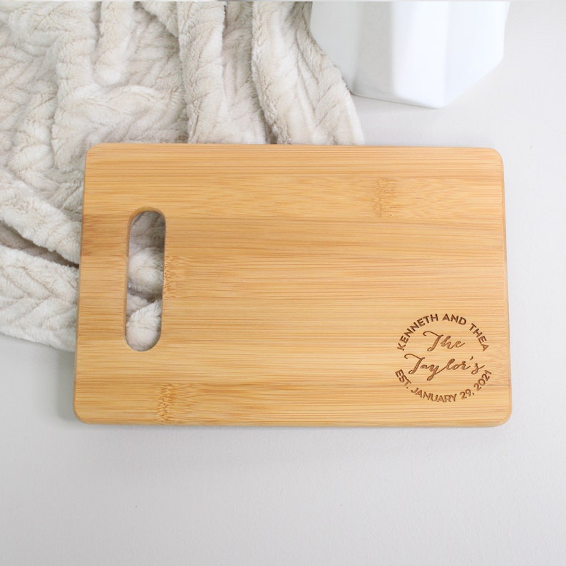 May include: A natural bamboo cutting board with a handle and a personalized inscription that reads "Kenneth and Thea The Taylor's Est. January 29, 2021".