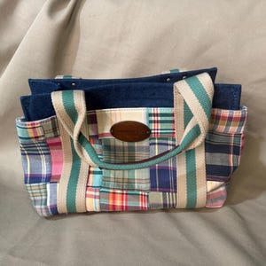 May include: A patchwork tote bag with a denim top and teal and white striped handles. The bag features a variety of plaid patterns in pink, blue, green, and yellow. A small, oval, brown leather tag is centered on the front.