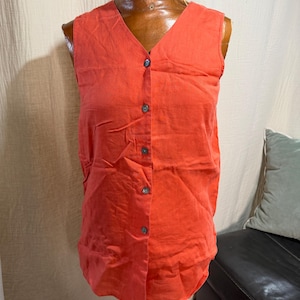 May include: A sleeveless, V-neck button-down top in a vibrant coral-orange color. The garment features a front placket with five dark, circular buttons and a straight hemline, displayed on a brown mannequin torso against a neutral backdrop.