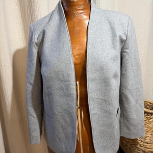 May include: A light gray blazer with a collarless, open-front design and long sleeves. The jacket features a textured fabric finish, straight hemline, and two side pockets, displayed on a brown mannequin torso.