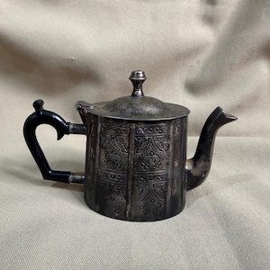 May include: Antique silver-colored teapot with a black handle. The teapot has a lid and a spout. The body of the teapot is decorated with an embossed pattern. The handle is curved and the spout is angled.