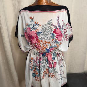 May include: A white dress with a floral print featuring pink, orange, and blue flowers. The dress has a black neckline and hem, with a gathered waist and short sleeves. The dress is displayed on a mannequin.