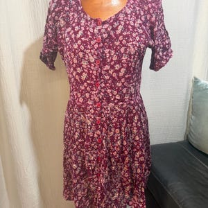 May include: A burgundy floral print dress with short sleeves and a button-down front. The dress features a scoop neckline and a fitted bodice that flows into a flared skirt. The fabric is covered in small, delicate white and pink flowers.