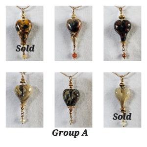 Puede incluir: Seis colgantes de vidrio en forma de corazón en cadenas doradas. Los colgantes son de varios colores y patrones, incluyendo marrón, dorado, negro y blanco. Algunos colgantes tienen una pequeña cuenta en la parte inferior. El texto "Sold" aparece en dos de los colgantes.
