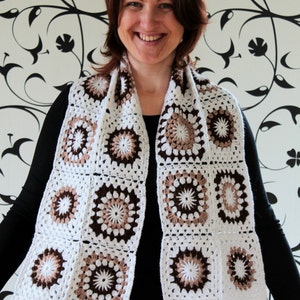 May include: A white crocheted scarf with brown and beige accents. The scarf is made of squares with a circular pattern in the center.