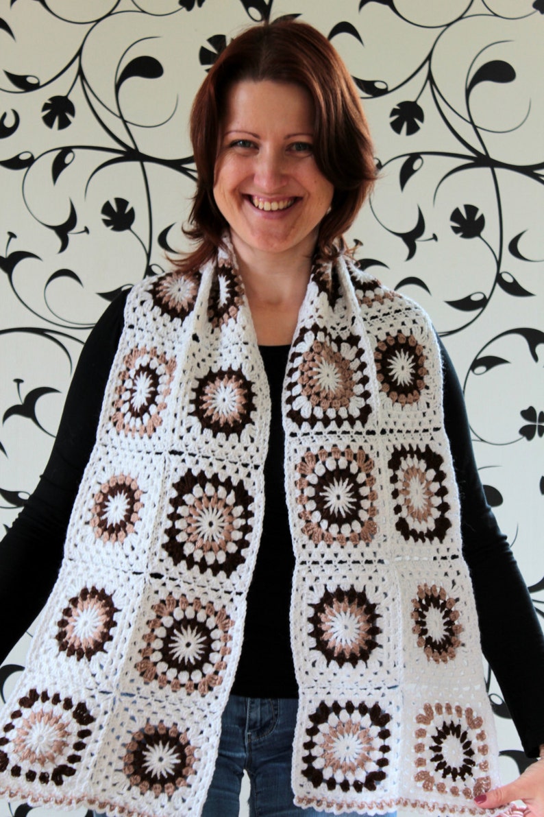 May include: A white crocheted scarf with brown and beige accents. The scarf is made of squares with a circular pattern in the center.