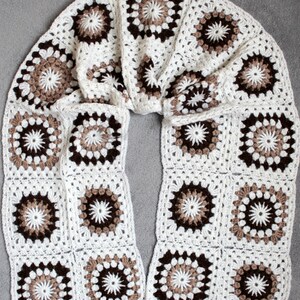 May include: A white crocheted scarf with brown and beige granny squares. The scarf is folded in half and laid flat on a gray surface.