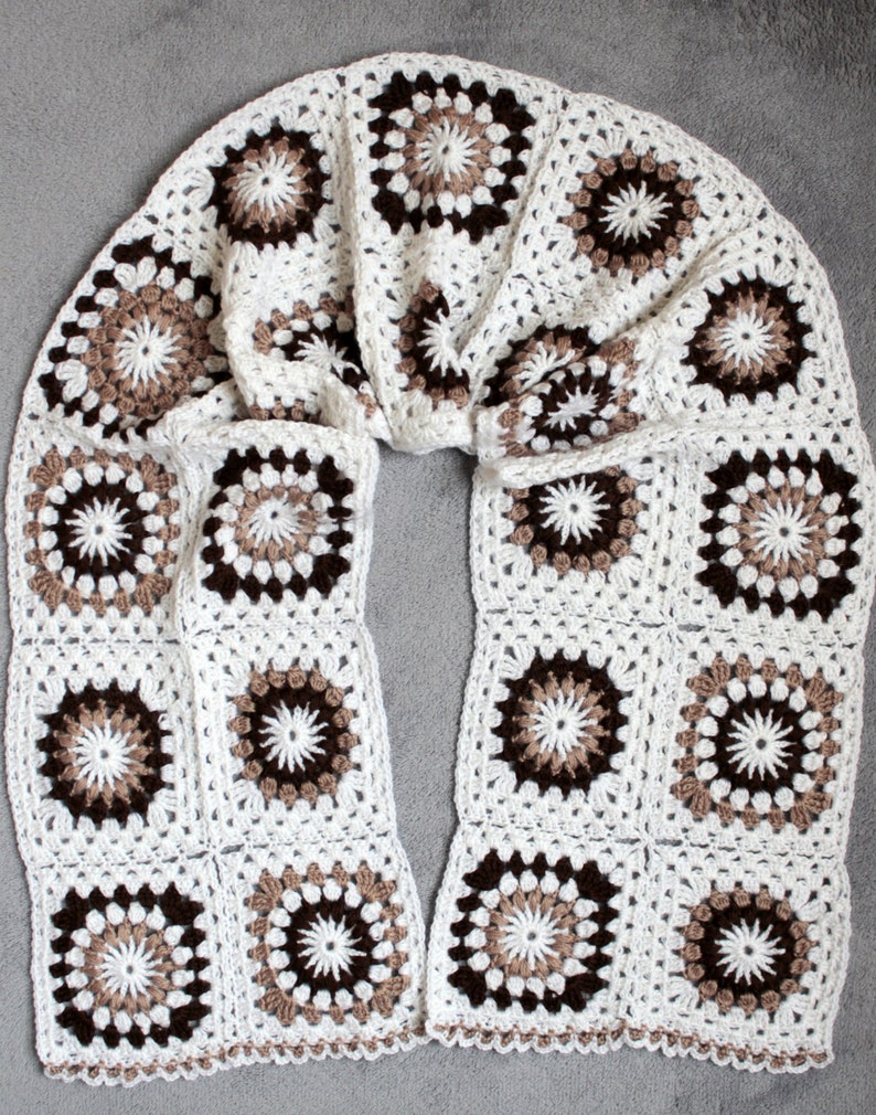 May include: A white crocheted scarf with brown and beige granny squares. The scarf is folded in half and laid flat on a gray surface.