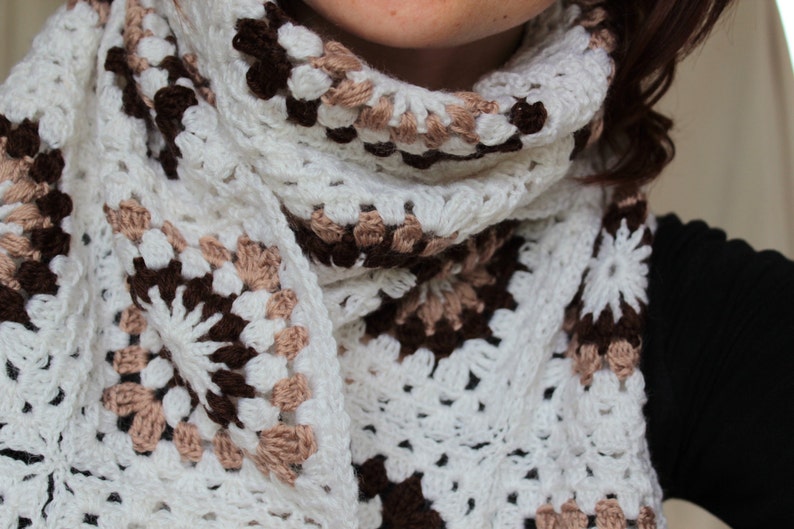 May include: A white crocheted scarf with brown and beige accents. The scarf is made of a delicate, lacy yarn and has a fringed edge.