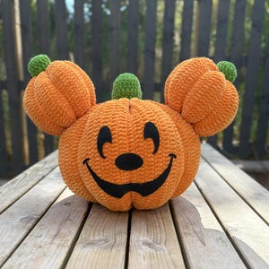 May include: A handmade, orange crochet pumpkin with a smiling face and green stem. The pumpkin has two smaller pumpkins on each side, resembling ears. Ideal for Halloween or autumn decorations.