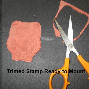 May include: A red rubber stamp in the shape of an owl, with intricate details, next to orange-handled scissors and a trimmed stamp. The text "Trimmed Stamp Ready to Mount" is visible. The items are on a dark surface.