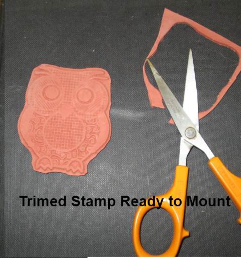 May include: A red rubber stamp in the shape of an owl, with intricate details, next to orange-handled scissors and a trimmed stamp. The text "Trimmed Stamp Ready to Mount" is visible. The items are on a dark surface.