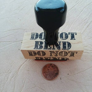 May include: A wooden stamp with the words "DO NOT BEND" in black, with a black handle. A copper penny is next to the stamp. The stamp is likely used for marking packages or documents.