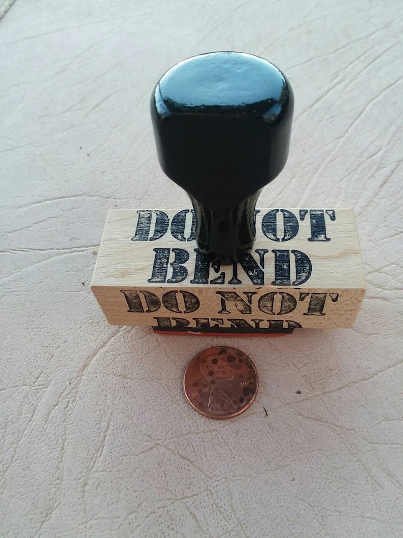 May include: A wooden stamp with the words "DO NOT BEND" in black, with a black handle. A copper penny is next to the stamp. The stamp is likely used for marking packages or documents.