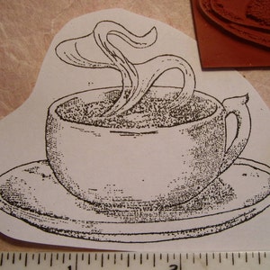 May include: Black and white line art of a coffee cup on a saucer, with steam rising from the cup. The image is a stamp, with a penny and a ruler in the background. The cup has a handle and sits on a round saucer.