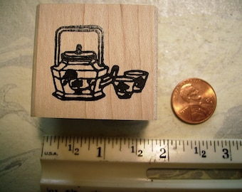 May include: A wooden stamp with a black illustration of a teapot and two cups. The stamp is on a light-coloured wooden block. A penny and a ruler are next to the stamp.