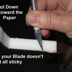 May include: A hand holding a craft knife cutting white foam. Text in the image reads "Cut Down toward the Paper" and "So your Blade doesn't get all sticky". The knife blade is angled downward, suggesting a specific cutting technique.