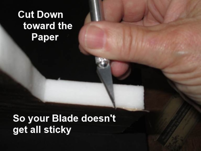 May include: A hand holding a craft knife cutting white foam. Text in the image reads "Cut Down toward the Paper" and "So your Blade doesn't get all sticky". The knife blade is angled downward, suggesting a specific cutting technique.