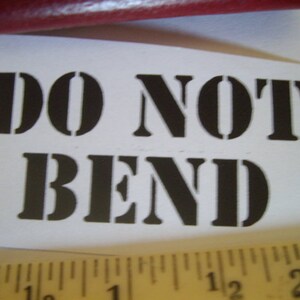 May include: A white paper with the words "DO NOT BEND" printed in bold, black stencil font. A wooden ruler with inch markings is at the bottom of the paper. A red object is at the top.