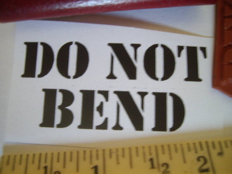 May include: A white paper with the words "DO NOT BEND" printed in bold, black stencil font. A wooden ruler with inch markings is at the bottom of the paper. A red object is at the top.