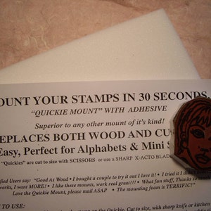 May include: A rubber stamp with a woman's face, in shades of red and brown, rests on a white foam pad. Text on a white paper reads "Mount Your Stamps in 30 Seconds" and "Quickie Mount".