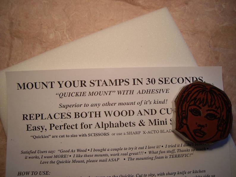 May include: A rubber stamp with a woman's face, in shades of red and brown, rests on a white foam pad. Text on a white paper reads "Mount Your Stamps in 30 Seconds" and "Quickie Mount".