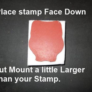 May include: A red stamp on a white square, with the text "Place stamp Face Down" and "Cut Mount a little Larger Than your Stamp." The stamp is on a black surface. The stamp has a unique shape.