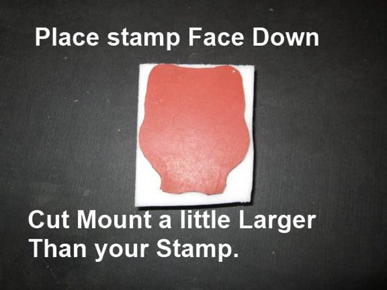 May include: A red stamp on a white square, with the text "Place stamp Face Down" and "Cut Mount a little Larger Than your Stamp." The stamp is on a black surface. The stamp has a unique shape.