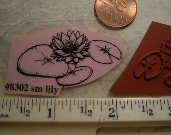May include: A pink rubber stamp featuring a black illustration of a water lily and lily pads. The text "#8302 sm lily" is printed on the stamp. A penny and a ruler are also in the image.