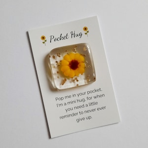 May include: A square clear resin token featuring a yellow sunflower with a dark center and gold flakes, attached to a white card. The card displays two small sunflower illustrations, the text Pocket Hug, and a message about a mini hug reminder.