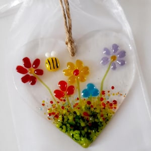 May include: A heart-shaped glass decoration with a floral design. The ornament features red, yellow, and purple flowers, a yellow and black bee, and a blue cloud. It hangs from a natural twine string and is presented in a sheer white bag.