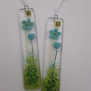 May include: Two clear glass bookmarks with floral designs. Each bookmark features a turquoise flower, a turquoise heart, and a yellow and black bee. Green and blue accents create a grassy base. White ribbon is attached for hanging.