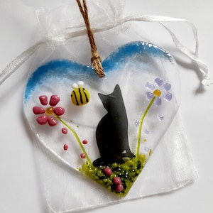 May include: A heart-shaped glass ornament with a black cat silhouette on green grass, pink berries, and flowers. It has a blue and white border, a yellow and black bee, and hangs from a jute string. The ornament is presented in a white organza bag.