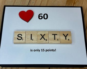 May include: A rectangular greeting card with a white background and black border. It features a red heart icon, the number 60, and wooden letter tiles spelling SIXTY. Below the tiles, the text reads: is only 15 points! The design is clean.