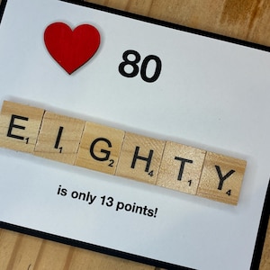 May include: A rectangular greeting card with a white background and black border. It features a red heart icon, the number 80, and the word EIGHTY spelled out with wooden letter tiles. Below the tiles, the text reads is only 13 points!