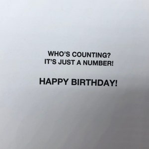 Op de afbeelding: Een witte wenskaart met gecentreerde zwarte tekst op de voorzijde. De tekst luidt: WHO'S COUNTING? IT'S JUST A NUMBER! HAPPY BIRTHDAY! De letters zijn in een strak, vet, schreefloos lettertype, gepresenteerd tegen een effen witte achtergrond.