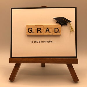 May include: A white card with black trim on a wooden easel. The card has a graduation cap and the word "GRAD" spelled out with Scrabble tiles. The text "is only 6 in scrabble....." is written below the tiles.