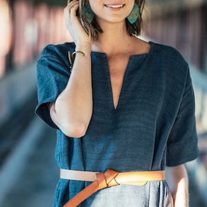 May include: A woman wearing a blue linen dress with a brown leather belt. She is wearing turquoise earrings and a gold bracelet.