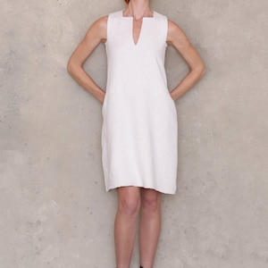 May include: A woman wearing a white sleeveless dress and black ankle boots. She is standing in front of a textured wall and wearing sunglasses.