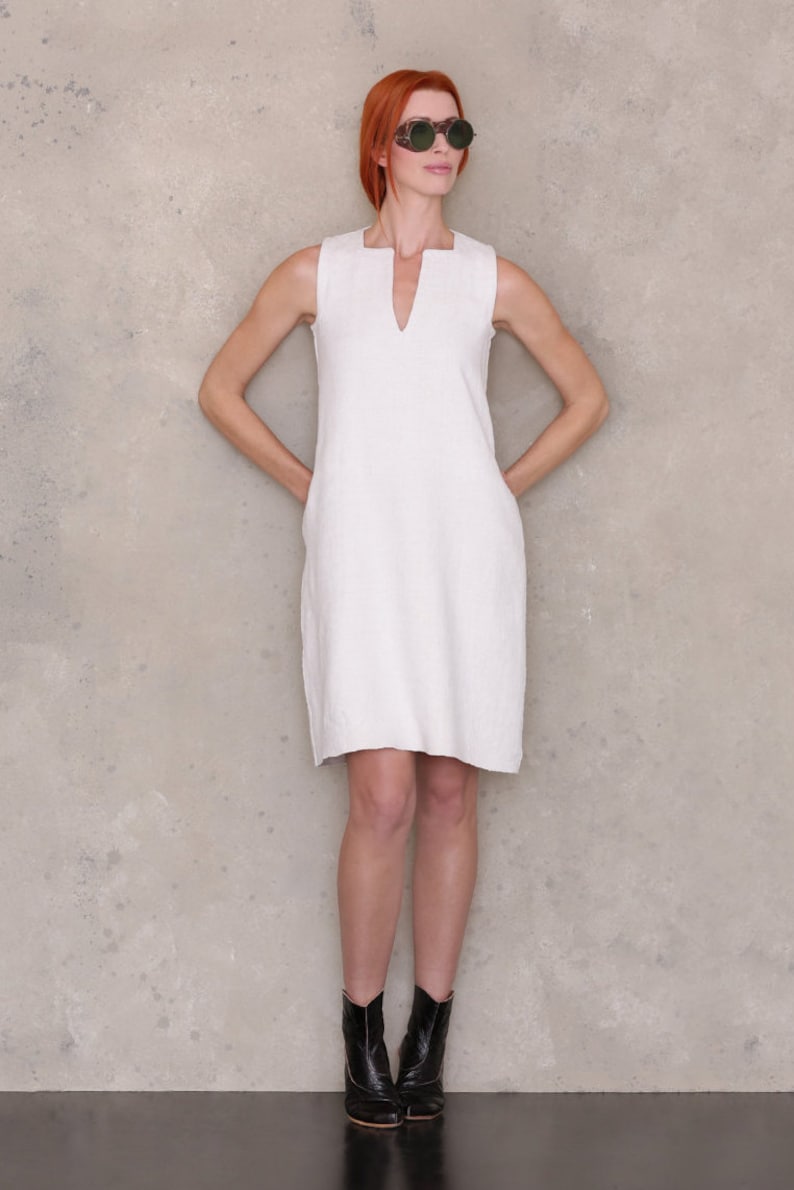 May include: A woman wearing a white sleeveless dress and black ankle boots. She is standing in front of a textured wall and wearing sunglasses.