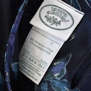 K&ouml;nnte beinhalten: Ein wei&szlig;es Etikett mit schwarzem Text, "laura ashley", auf einem dunkelblauen Stoff mit Blumenmuster. Das Etikett listet die Stoffzusammensetzung als 89% Viskose/Rayon/Viskose und 11% Wolle/Wolle/Wol/Wolle auf. Das Etikett listet auch die Gr&ouml;&szlig;e als UK 12, EUR 38, USA 8 und die RN-Nummer 73785 auf.