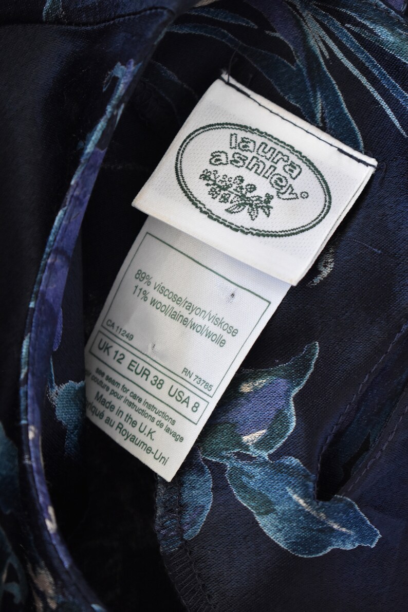 K&ouml;nnte beinhalten: Ein wei&szlig;es Etikett mit schwarzem Text, "laura ashley", auf einem dunkelblauen Stoff mit Blumenmuster. Das Etikett listet die Stoffzusammensetzung als 89% Viskose/Rayon/Viskose und 11% Wolle/Wolle/Wol/Wolle auf. Das Etikett listet auch die Gr&ouml;&szlig;e als UK 12, EUR 38, USA 8 und die RN-Nummer 73785 auf.