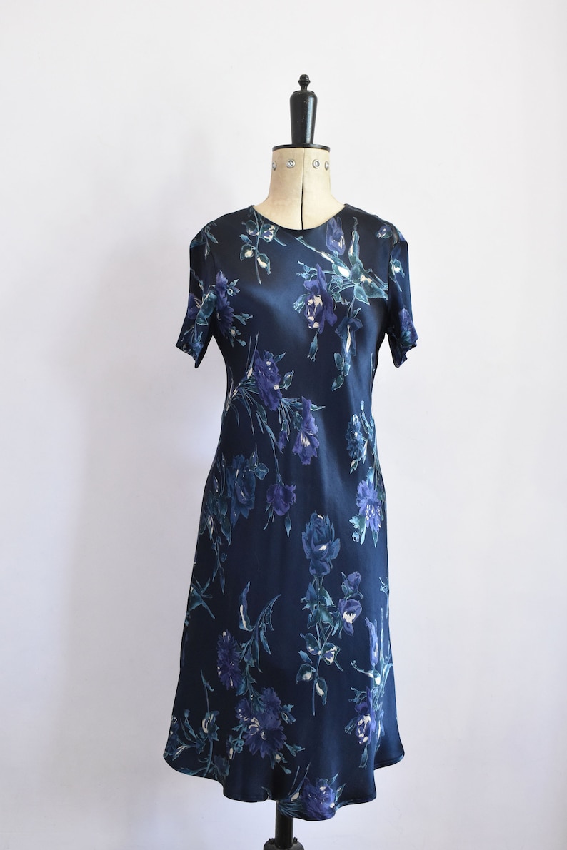 K&ouml;nnte beinhalten: Ein dunkelblaues Satin-Kleid mit einem Blumendruck in Blau- und Gr&uuml;nt&ouml;nen. Das Kleid hat kurze &Auml;rmel und ein tailliertes Oberteil.
