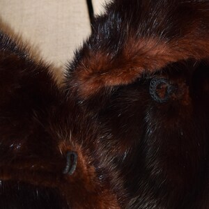 May include: Close-up of a luxurious fur coat with a rich, dark brown color and a contrasting reddish-brown trim. The fur appears soft and dense, with visible stitching and button details. The coat's texture and color suggest a vintage or high-end garment.