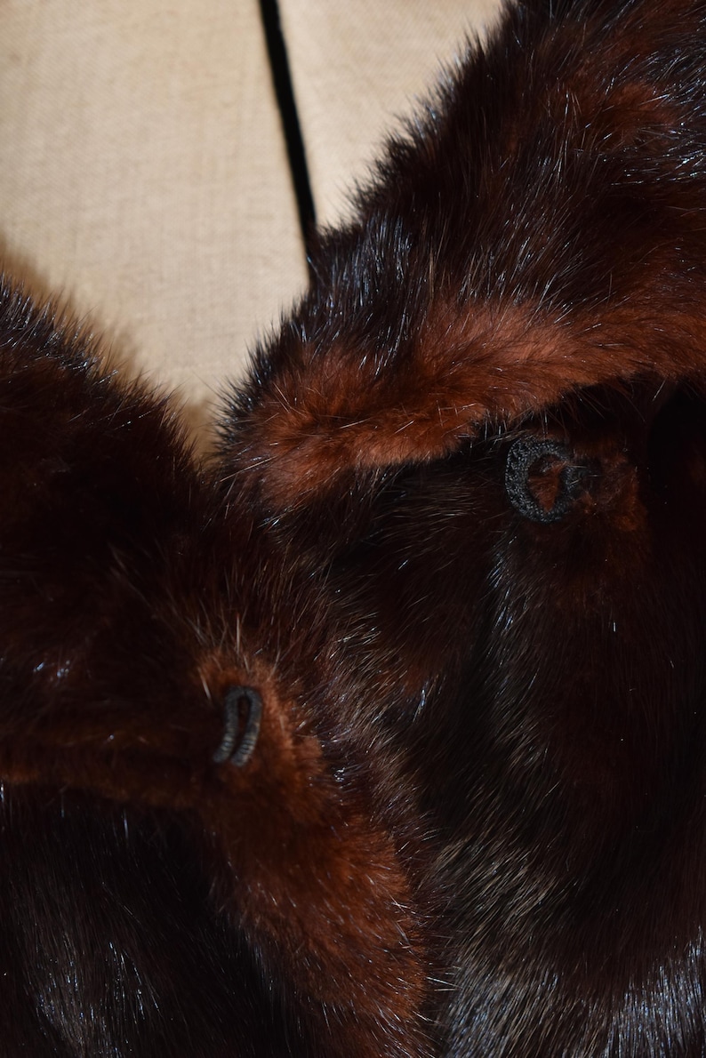 May include: Close-up of a luxurious fur coat with a rich, dark brown color and a contrasting reddish-brown trim. The fur appears soft and dense, with visible stitching and button details. The coat's texture and color suggest a vintage or high-end garment.