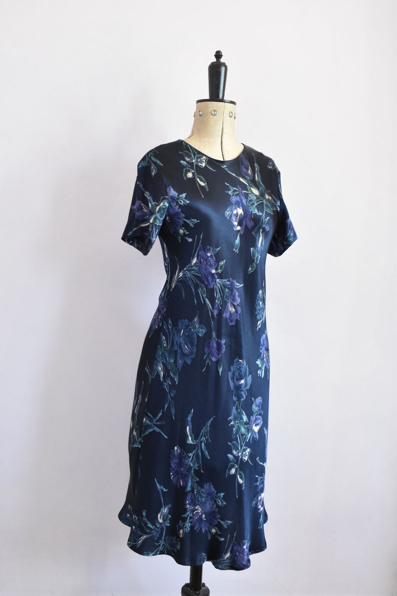 K&ouml;nnte beinhalten: Ein dunkelblaues Satin-Kleid mit einem Blumendruck mit blauen und wei&szlig;en Blumen. Das Kleid hat kurze &Auml;rmel und eine figurbetonte Silhouette.