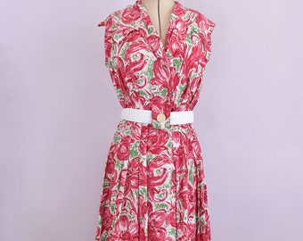 Könnte beinhalten: Ein ärmelloses Kleid mit weißem Kragen und einem Blumendruck in Rosa- und Grüntönen. Das Kleid hat einen weißen Gürtel mit einem goldenen Knopf.