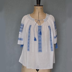May include: White blouse with blue floral embroidery. The blouse has a V-neck with a drawstring and tassels, and short sleeves. The embroidery is detailed and covers the neckline, sleeves, and front panels.