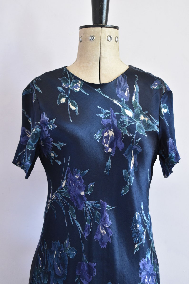 K&ouml;nnte beinhalten: Ein dunkelblaues Satin-Kleid mit einem Blumendruck mit blauen und wei&szlig;en Blumen. Das Kleid hat kurze &Auml;rmel und einen runden Ausschnitt.